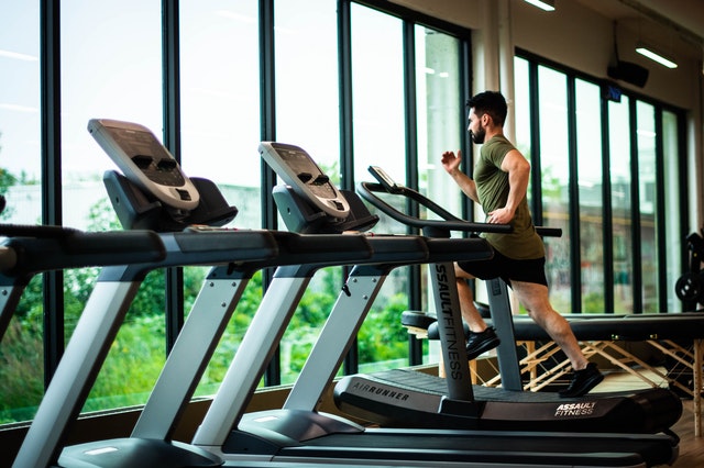 A man runs on a treadmill in a gym in front of some windows